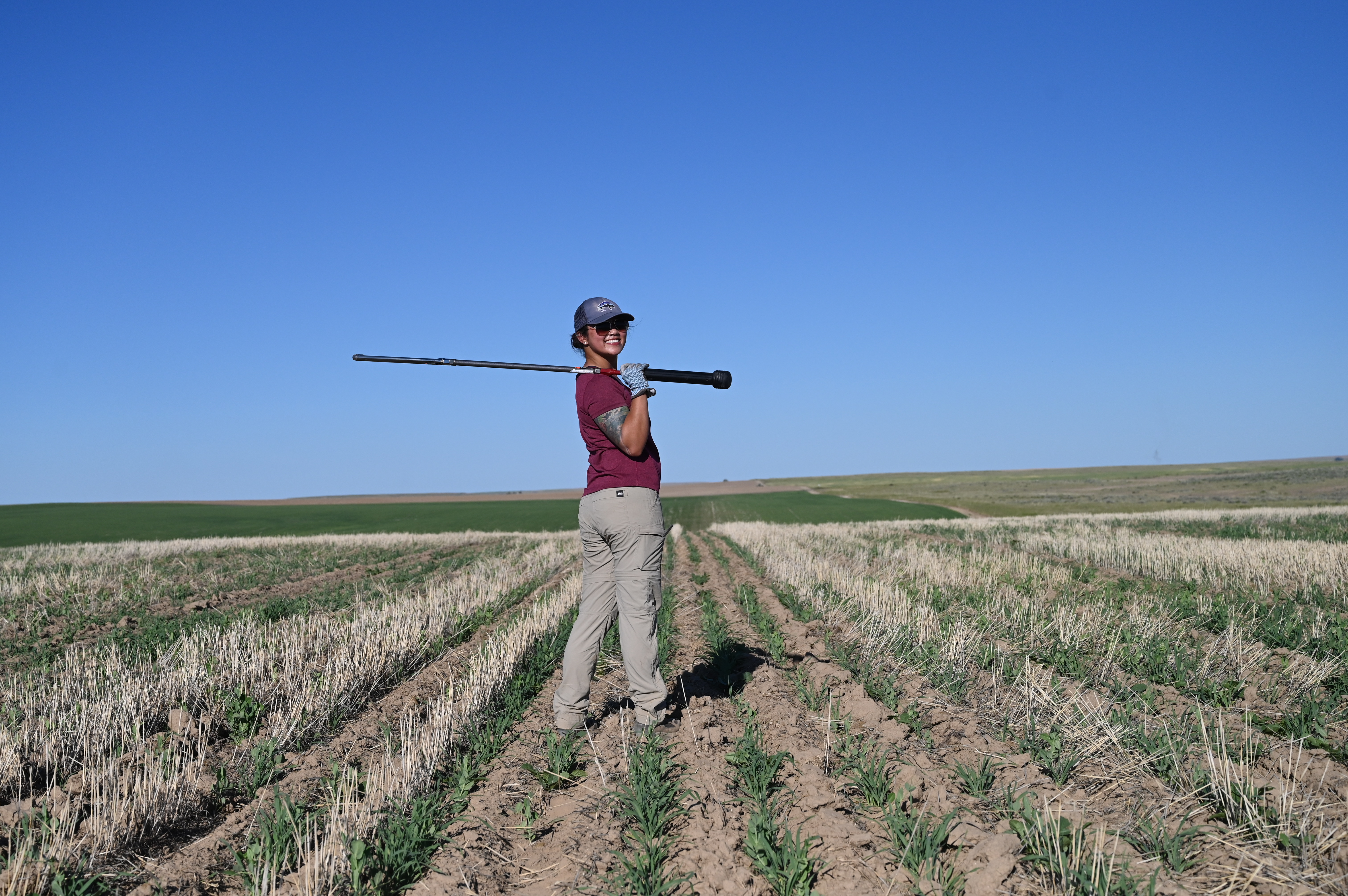 Jadey standing in a field of wheat stubble holding a 3 foot long soil sampling probe over her shoulder.